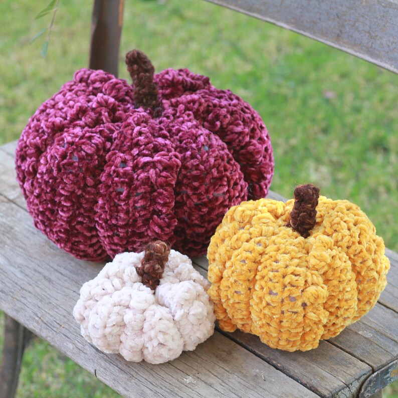 May include: Three crocheted pumpkins in different sizes and colors. The largest pumpkin is a deep burgundy color, the medium pumpkin is a light beige color, and the smallest pumpkin is a bright yellow color.