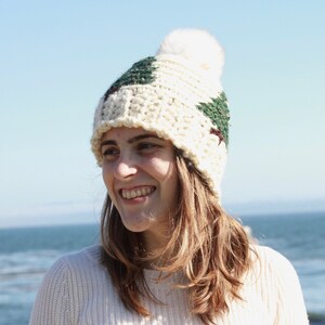 May include: A woman wearing a white crocheted beanie with a green and brown pattern and a white pom pom. The beanie is pulled down over her ears and she is smiling.