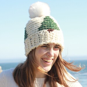May include: A white crocheted beanie with a green and brown tree design and a white pom pom on top.
