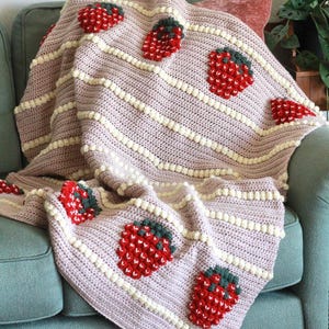 K&ouml;nnte beinhalten: Eine rosa-wei&szlig;e H&auml;keldecke mit Erdbeer-Design. Die Decke ist gefaltet und &uuml;ber ein t&uuml;rkisfarbenes Sofa gelegt.