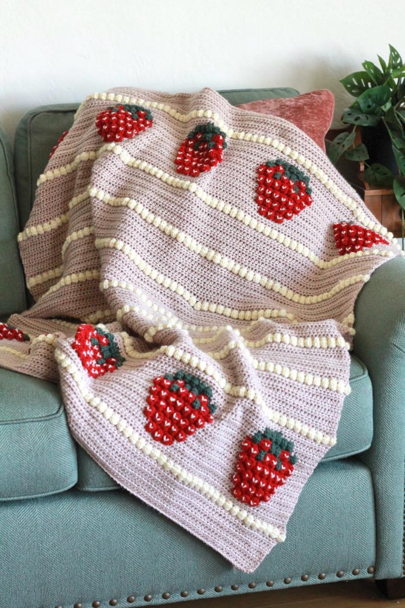 K&ouml;nnte beinhalten: Eine rosa-wei&szlig;e H&auml;keldecke mit Erdbeer-Design. Die Decke ist gefaltet und &uuml;ber ein t&uuml;rkisfarbenes Sofa gelegt.