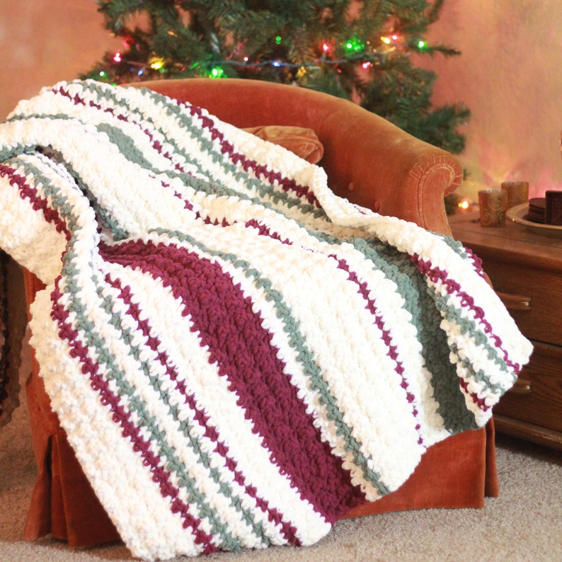 May include: A white, burgundy, and green striped crocheted throw blanket draped over an orange armchair. The blanket is made with a chunky yarn and has a textured, fluffy look.