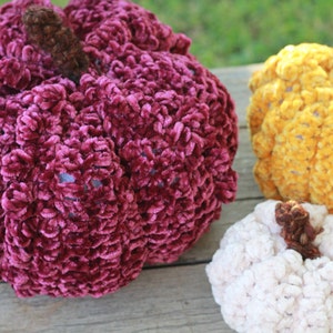 May include: Three crocheted pumpkins in different colors: burgundy, yellow, and white. The pumpkins are made with a fuzzy yarn and have a textured surface.