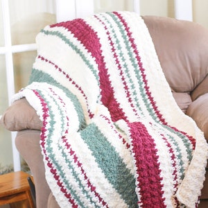 May include: A striped crocheted throw blanket in shades of white, burgundy, and green. The blanket is draped over a brown armchair.