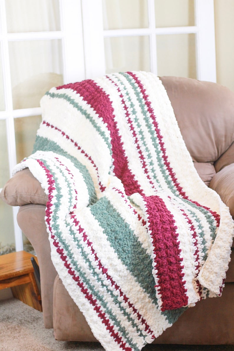 May include: A striped crocheted throw blanket in shades of white, burgundy, and green. The blanket is draped over a brown armchair.