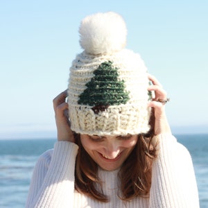 May include: A white crocheted beanie with a green and brown tree design and a white pom pom on top.