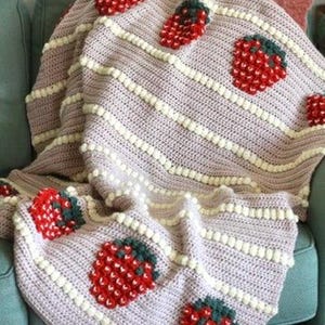 K&ouml;nnte beinhalten: Eine geh&auml;kelte Wolldecke mit rosa Hintergrund und wei&szlig;en Streifen. Die Decke zeigt ein sich wiederholendes Muster aus roten und gr&uuml;nen Erdbeeren.