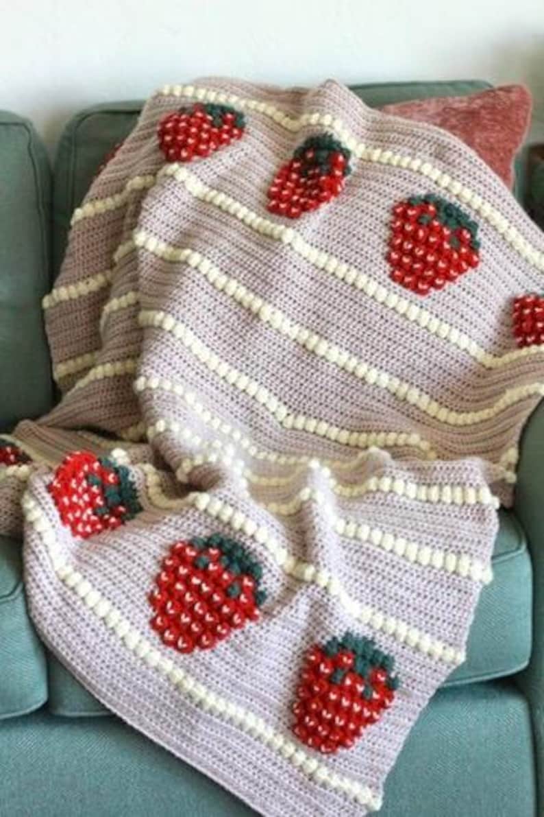 K&ouml;nnte beinhalten: Eine geh&auml;kelte Wolldecke mit rosa Hintergrund und wei&szlig;en Streifen. Die Decke zeigt ein sich wiederholendes Muster aus roten und gr&uuml;nen Erdbeeren.