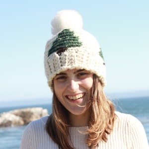 May include: A white crocheted beanie with a green and brown tree design and a white pom pom on top. The beanie is worn by a person with long brown hair and a white sweater.