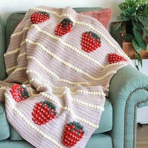K&ouml;nnte beinhalten: Eine geh&auml;kelte Wolldecke mit einem rosa-wei&szlig;en Streifenmuster und gestickten Erdbeeren. Die Decke liegt auf einem t&uuml;rkisfarbenen Sofa.