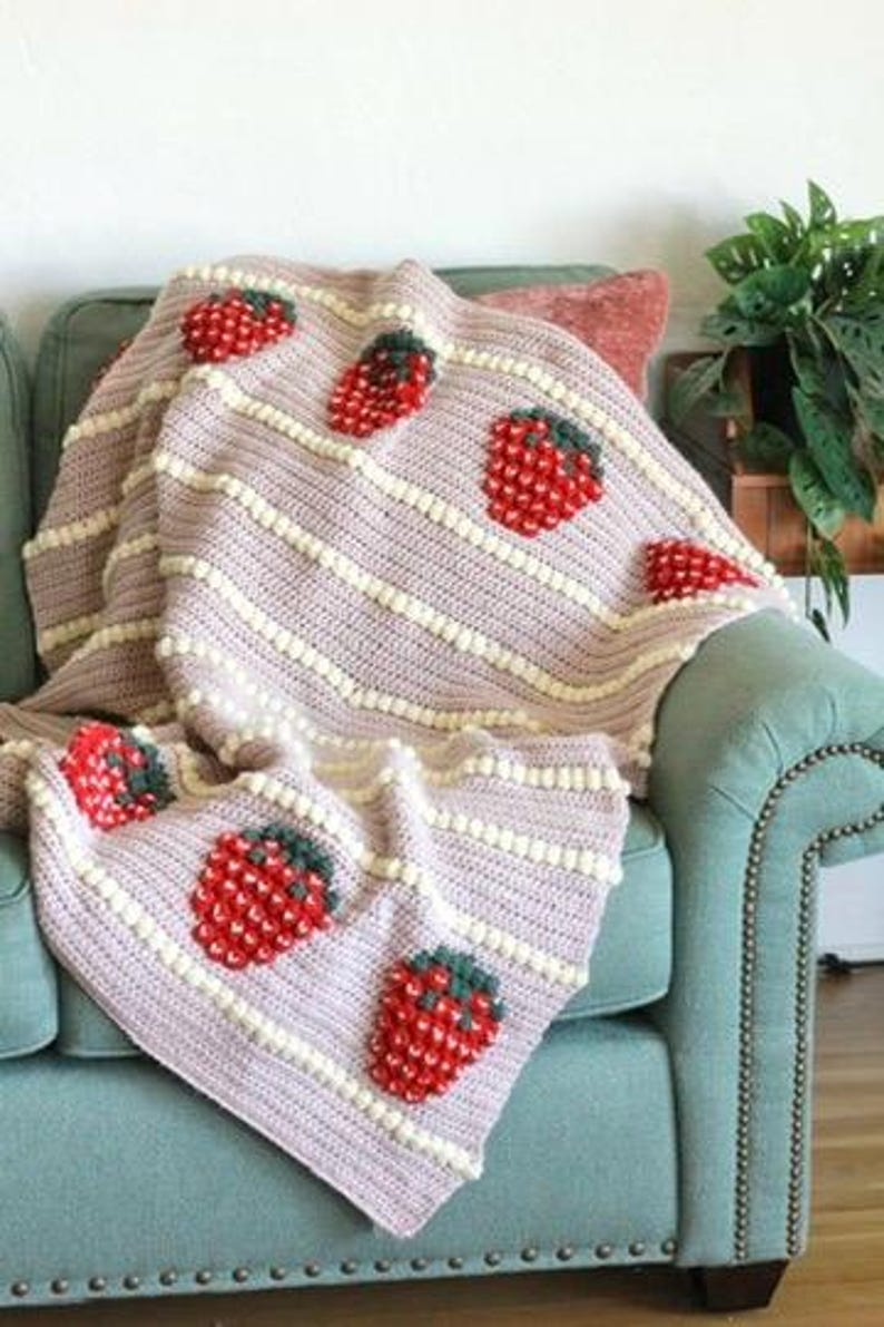 K&ouml;nnte beinhalten: Eine geh&auml;kelte Wolldecke mit einem rosa-wei&szlig;en Streifenmuster und gestickten Erdbeeren. Die Decke liegt auf einem t&uuml;rkisfarbenen Sofa.