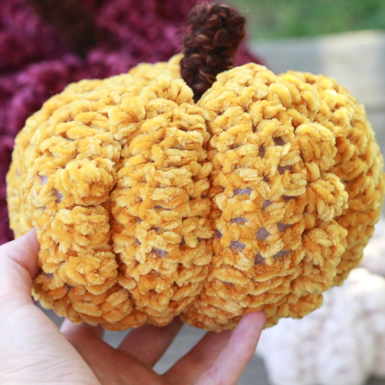 May include: A crocheted pumpkin in a bright yellow color with a brown stem. The pumpkin is made with a fuzzy yarn that gives it a soft, plush texture.