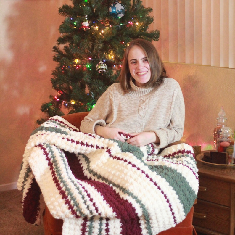 May include: A person sitting in a chair with a white, green, and burgundy striped crocheted blanket. The person is smiling and looking at the camera. There is a Christmas tree in the background.