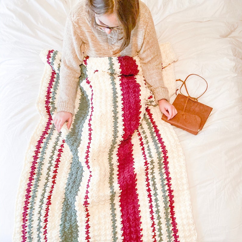 May include: A white, red, and green striped crocheted blanket with a textured, puffy design. The blanket is folded and draped over a person sitting on a white bed.