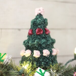 Puede incluir: Un &aacute;rbol de Navidad de crochet verde con adornos de fieltro rosa y burdeos. El &aacute;rbol est&aacute; sentado en una guirnalda verde con peque&ntilde;os regalos envueltos y luces blancas.