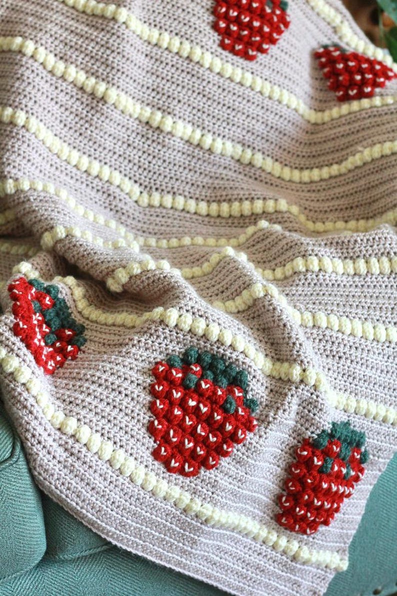 K&ouml;nnte beinhalten: Eine rosa geh&auml;kelte Decke mit wei&szlig;em Rand und Erdbeer-Designs. Die Erdbeeren sind aus rotem, wei&szlig;em und gr&uuml;nem Garn gefertigt.
