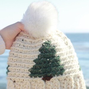 May include: White crochet beanie with a green pine tree design and a white pom pom. The beanie is made with a sparkly yarn. The text "Pinewood Beanie Crochet Winter Hat Pattern by ECLAIReMAKERY.COM" is at the bottom of the image.