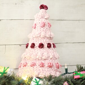 Puede incluir: Un adorno de &aacute;rbol de Navidad de crochet rosa y burdeos con textura de terciopelo. El &aacute;rbol est&aacute; decorado con peque&ntilde;as pompones rosas y burdeos.