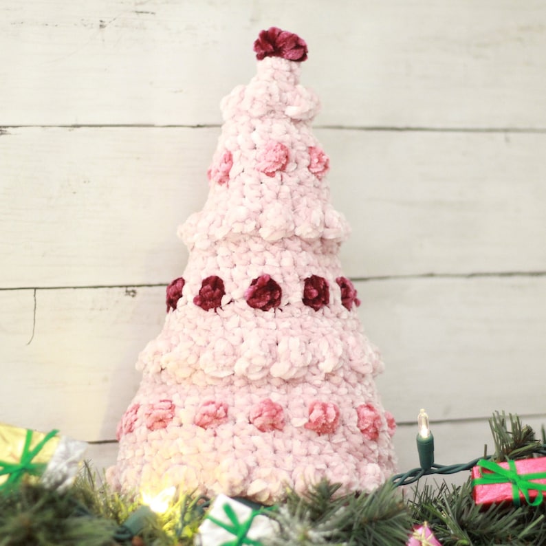 Puede incluir: Un adorno de &aacute;rbol de Navidad de crochet rosa y burdeos con textura de terciopelo. El &aacute;rbol est&aacute; decorado con peque&ntilde;as pompones rosas y burdeos.