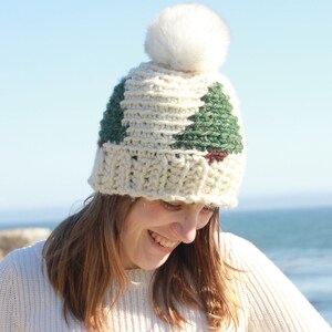 May include: A white crocheted beanie with a green and brown tree design and a white pom pom on top. The beanie is worn by a person with brown hair and is smiling.