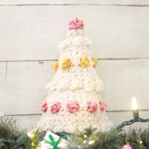 Puede incluir: Un &aacute;rbol de Navidad de crochet blanco decorado con pompones rosas y amarillos. El &aacute;rbol est&aacute; sobre un fondo de madera blanca con una cadena de luces.