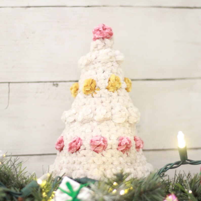 Puede incluir: Un &aacute;rbol de Navidad de crochet blanco decorado con pompones rosas y amarillos. El &aacute;rbol est&aacute; sobre un fondo de madera blanca con una cadena de luces.