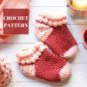 May include: A pair of crocheted slippers in pink and red with a ruffled edge. The slippers are on a white wooden surface. The text "CROCHET PATTERN" is visible in the image.