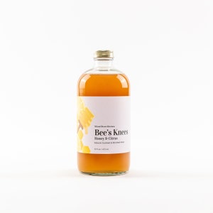 May include: A glass bottle of Bee's Knees Honey & Citrus natural cocktail and mocktail mixer. The bottle is filled with a light amber liquid and has a white label with black text. The label features a graphic of a honeycomb and a lemon.