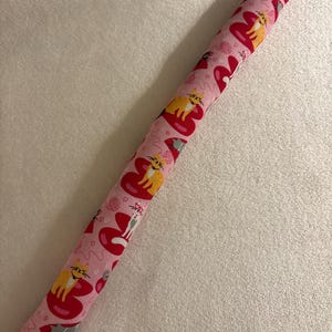 May include: A pink cat toy with a repeating pattern of cats on red hearts. The cats are yellow and white, and the hearts have a red outline. The toy is long and cylindrical, perfect for cat play.