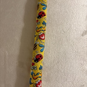 May include: A yellow fabric tube with a repeating pattern of cartoon insects, including ladybugs, bees, and smiling spiders. The insects are in red, blue, and black, with a whimsical design. The tube is likely a decorative item or a part of a larger craft project.