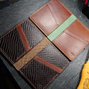 May include: Two leather wallets displayed open. One wallet features a dark brown snakeskin-textured leather with smooth brown accents and yellow stitching. The other wallet is smooth tan leather with a light green center panel and yellow stitching.