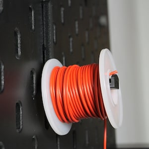 May include: A spool of bright orange electrical wire mounted on a black pegboard. The wire is neatly wound on a white plastic spool, with a small black clip securing it to the board. The background is blurred.