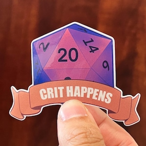 May include: A sticker featuring a D20 die with the number 20 prominently displayed. The die is pink and purple with blue accents. Below the die is a banner that reads "CRIT HAPPENS" in white text.