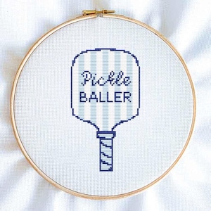 May include: Cross-stitch embroidery in a wooden hoop. The design features a pickleball paddle with the words "Pickle Baller" in dark blue. The paddle has light blue and white vertical stripes. The background is white.