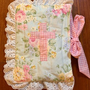 May include: A floral-patterned book cover with a pink gingham cross applique. The cover features a light blue background with pink, yellow, and white flowers. It has a white eyelet lace trim and a pink gingham ribbon tie.