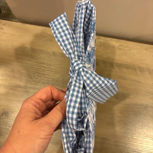 Op de afbeelding: Een blauw-wit geruite stof die in een strik is gebonden. De stof is om een wit voorwerp gewikkeld. De stof heeft een bloemenpatroon. Het voorwerp wordt door een persoon vastgehouden.
