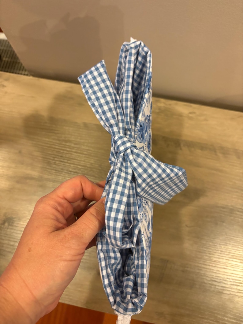 Op de afbeelding: Een blauw-wit geruite stof die in een strik is gebonden. De stof is om een wit voorwerp gewikkeld. De stof heeft een bloemenpatroon. Het voorwerp wordt door een persoon vastgehouden.
