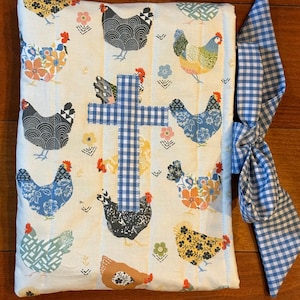 May include: A quilted book cover featuring a repeating pattern of colorful chickens on a cream background with vertical stripes. A blue and white gingham cross is centered on the cover. A blue and white gingham ribbon is tied to the side.