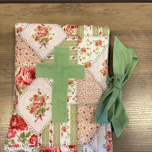 May include: A quilted Bible cover with a green cross applique. The cover features a patchwork design with floral patterns in red, pink, and white. A matching green fabric napkin is tied to the side.