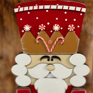 Peut inclure: Une figurine de casse-noisette en bois avec un chapeau rouge et blanc, une couronne brune avec des cannes en sucre et une barbe blanche. Le casse-noisette est peint avec des pois blancs et a un visage brun.