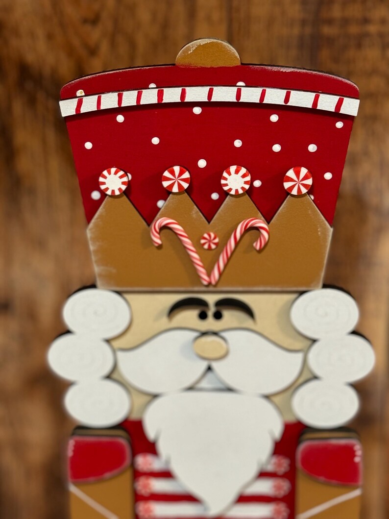 Peut inclure: Une figurine de casse-noisette en bois avec un chapeau rouge et blanc, une couronne brune avec des cannes en sucre et une barbe blanche. Le casse-noisette est peint avec des pois blancs et a un visage brun.