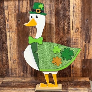 May include: A white goose wearing a green shamrock patterned coat, a green bow tie, and a green leprechaun hat with a gold buckle. The goose is standing on a wooden base.