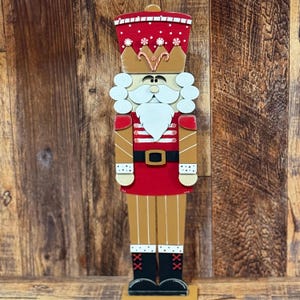 May include: A wooden nutcracker figurine with a red and white striped uniform, a gold crown, and a white beard. The nutcracker is standing on a wooden base.