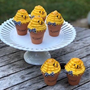 May include: Six yellow and brown cupcakes decorated with honey and bees, displayed on a white cake stand and a wooden table. The cupcakes are labeled "Hunny".
