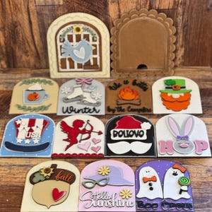 May include: A collection of decorative wooden cutouts with various seasonal and holiday themes. Designs include a bird in a window, a campfire, a leprechaun, and a snowman with the text "Doliovo". Other designs include a hat, acorn, and ghosts.