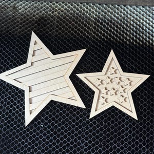 May include: Two wooden star cutouts. One star has horizontal lines, the other has smaller star cutouts. Both are light brown and sit on a black mesh surface.