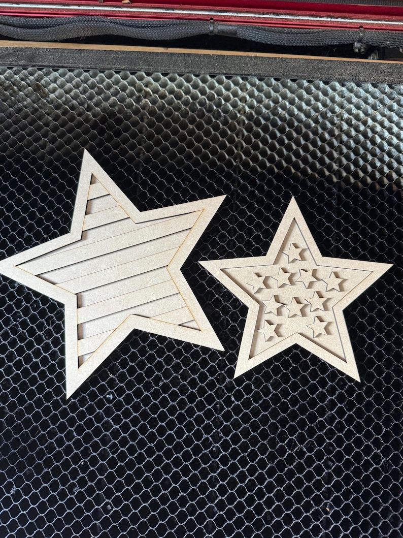 May include: Two wooden star cutouts. One star has horizontal lines, the other has smaller star cutouts. Both are light brown and sit on a black mesh surface.