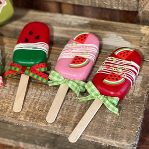 May include: Three watermelon popsicle-shaped decorations with wooden sticks and green and white gingham ribbon bows. The popsicles are red, pink, and green with white frosting and watermelon slices.