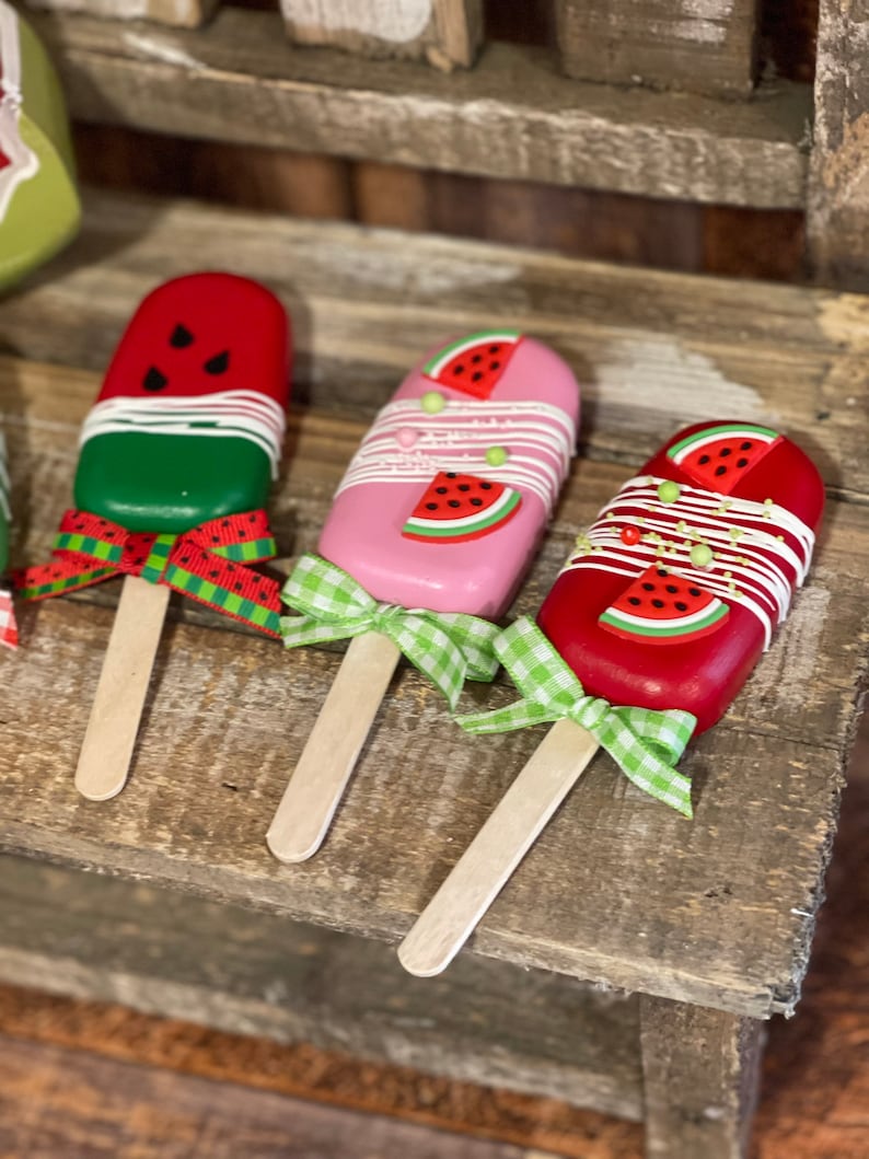 May include: Three watermelon popsicle-shaped decorations with wooden sticks and green and white gingham ribbon bows. The popsicles are red, pink, and green with white frosting and watermelon slices.