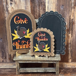May include: Thanksgiving-themed wooden signs with the words "Give Thanks" and a turkey illustration. The signs are in various shapes and sizes, with a brown and black color scheme. The signs are displayed on a small wooden bench.